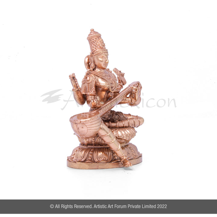 A bronze metal idol of Goddess Saraswati, depicted with four hands, sitting on a structure, with a veena musical instrument. The idol has a glossy finish.